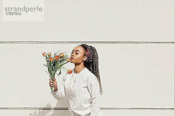 Junge Frau mit geschlossenen Augen riecht Blumen vor einer weißen Wand an einem sonnigen Tag