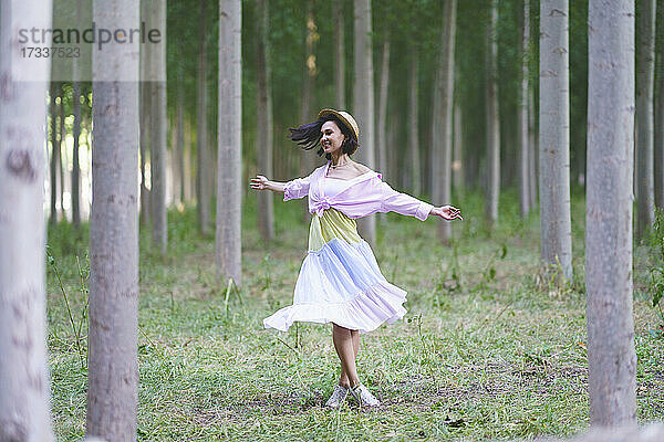 Lächelnde junge Frau tanzt mit ausgestreckten Armen im Wald