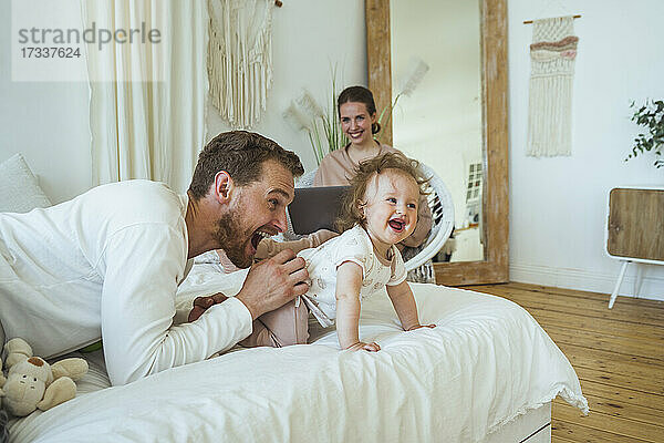 Lächelnde Frau schaut auf einen fröhlichen Mann  der mit seiner Tochter im Schlafzimmer spielt