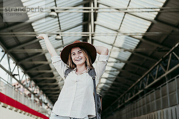 Fröhliche junge Passagierin mit Hut steht mit erhobenen Händen am Bahnhof