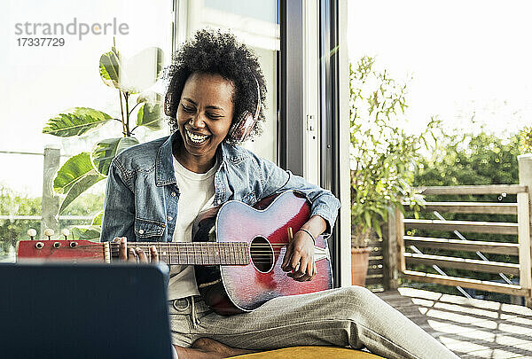 Fröhliche Frau mit Kopfhörern spielt Gitarre  während sie zu Hause mit einem digitalen Tablet lernt