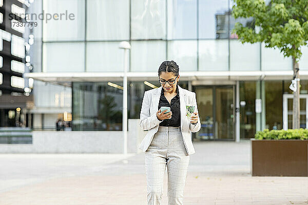Geschäftsfrau  die ein Smartphone benutzt  während sie vor einem Bürogebäude spazieren geht