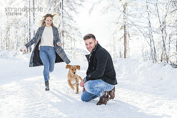 Mann hockt im Schnee  während seine Freundin im Urlaub mit dem Hund im Schnee läuft