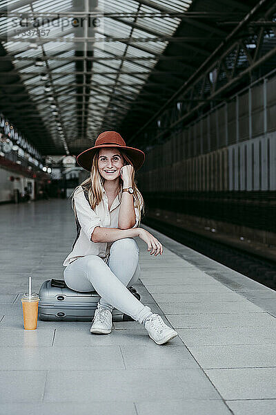Glückliche Frau mit Hut  die auf einem Gepäckstück auf dem Bahnsteig sitzt