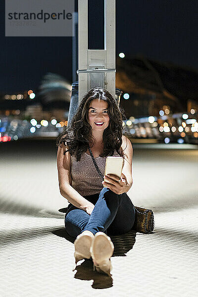 Lächelnde Frau mit Mobiltelefon auf dem Boden sitzend am Pier bei Nacht