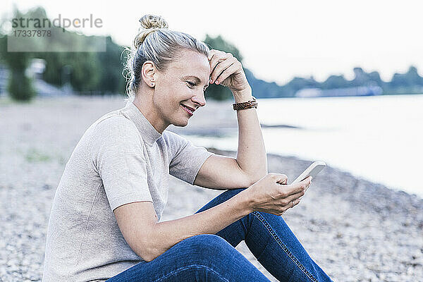 Frau  die am Flussufer sitzend ein Mobiltelefon benutzt