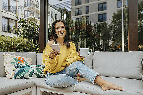 Frau lacht  während sie Tasse und Smartphone auf dem Sofa im Hinterhof hält
