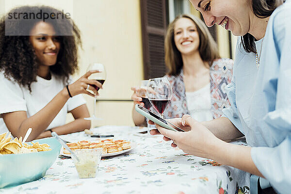 Lächelnde Frau  die ein Mobiltelefon benutzt  während sie mit Freunden auf der Terrasse sitzt