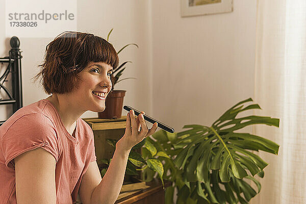 Lächelnde Frau  die eine Sprachnachricht über ihr Smartphone sendet  während sie zu Hause im Schlafzimmer sitzt