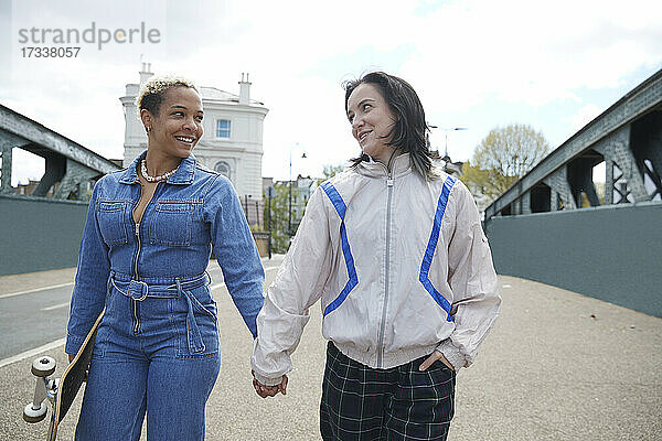 Lächelnde Freundinnen  die sich an den Händen halten und sich gegenseitig ansehen  während sie auf einer Brücke gehen