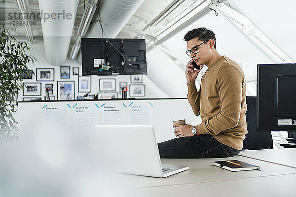 Geschäftsmann mit Laptop  der im Büro sitzt und mit seinem Smartphone telefoniert