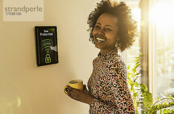 Fröhliche Frau mit Kaffeetasse  die neben einem Hausautomatisierungsgerät steht