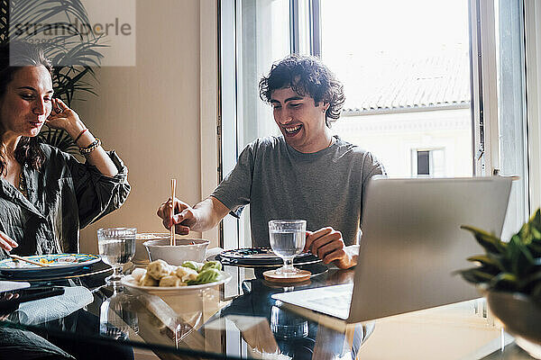 Lächelnder Mann isst Essen  während er neben einer Frau zu Hause sitzt