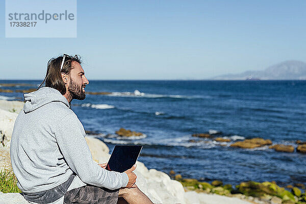 Lächelnder männlicher Freiberufler mit Blick auf das Meer  während er mit seinem Laptop am Strand sitzt