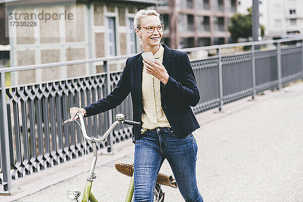 Lächelnde Geschäftsfrau mit Mobiltelefon in der Hand  während sie mit dem Fahrrad auf einer Brücke spazieren geht