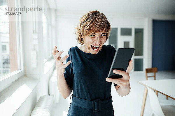 Aufgeregte Geschäftsfrau  die ein Mobiltelefon benutzt  während sie im Büro steht