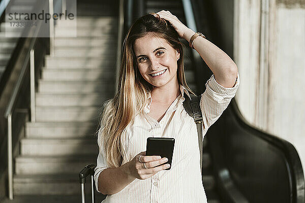 Glückliche junge Frau hält ihr Smartphone in der Hand  während sie mit der Hand im Haar steht