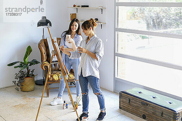 Eine Künstlerin malt auf einer Leinwand  während ihr Kollege im Atelier einen Becher hält