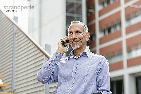Lächelnder Geschäftsmann  der mit seinem Handy telefoniert und sich auf ein Geländer stützt