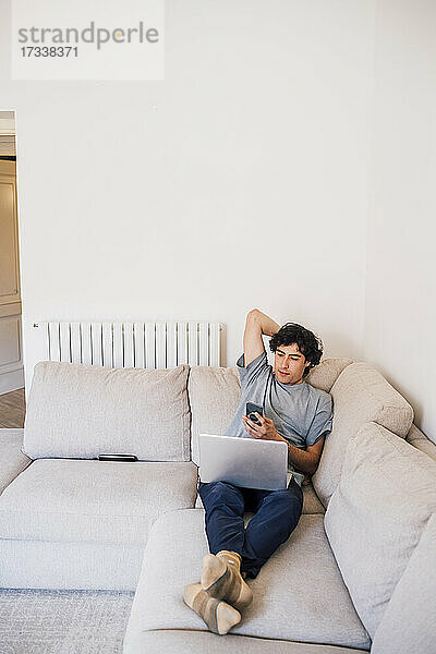 Mann  der ein Mobiltelefon benutzt  während er mit einem Laptop auf dem Sofa im Wohnzimmer sitzt