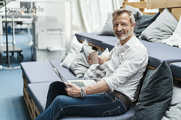 Lächelnde männliche Fachkraft mit Einwegbecher und Laptop auf dem Sofa sitzend