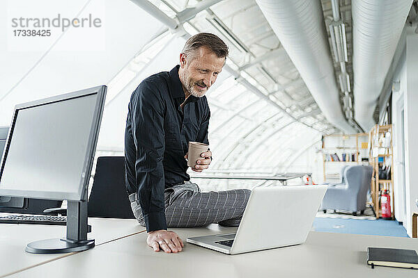 Männlicher Berufstätiger mit Kaffeetasse  der auf seinen Laptop schaut  während er im Büro sitzt