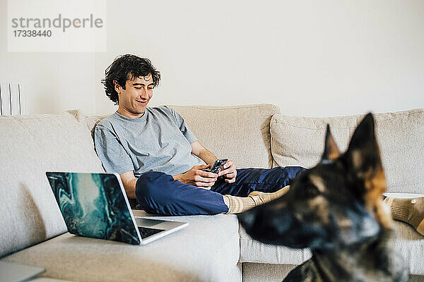 Lächelnder Mann  der ein Mobiltelefon benutzt  während er zu Hause auf dem Sofa am Laptop sitzt