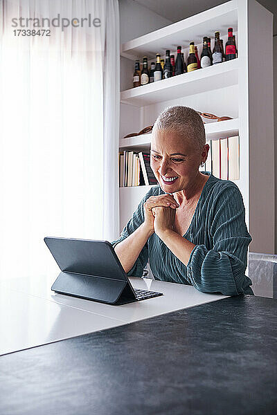 Lächelnde Frau schaut auf ein digitales Tablet  während sie im Wohnzimmer sitzt