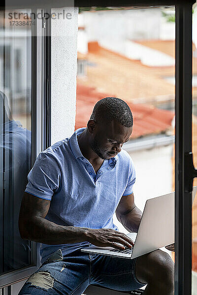 Mann benutzt Laptop  während er am Fenster seines Hauses sitzt