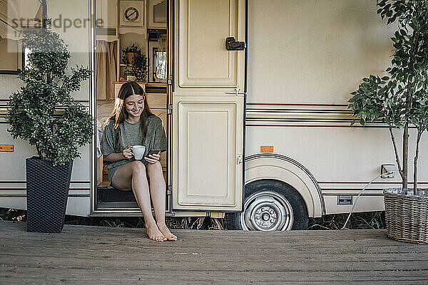 Lächelnde junge Frau  die eine SMS über ihr Smartphone schreibt  während sie an der Tür eines Wohnmobils sitzt