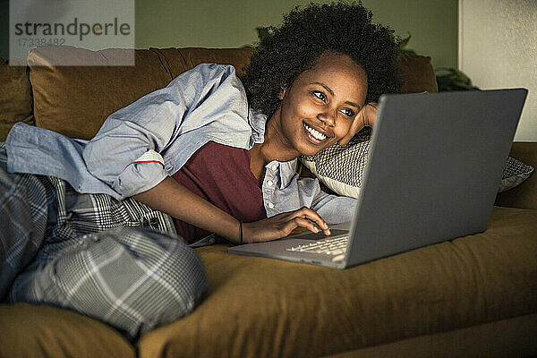 Junge Frau benutzt einen Laptop  während sie zu Hause auf dem Sofa liegt