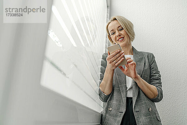 Blonde weibliche Berufstätige  die über ihr Smartphone am Fenster im Büro Textnachrichten verschickt