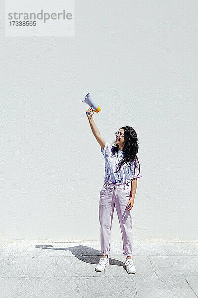 Junge Frau mit erhobener Hand  die ein Megaphon vor einer weißen Wand hält