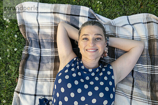 Lächelnde reife Frau mit Händen hinter dem Kopf auf einer Decke im Park liegend