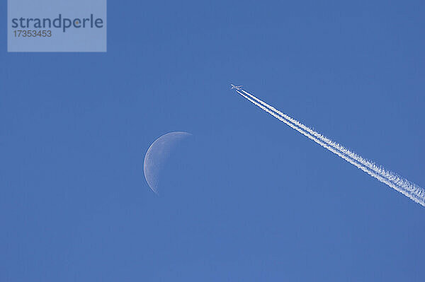 Flugzeug hinterlässt Kondensstreifen vor blauem Himmel mit abnehmendem Mond
