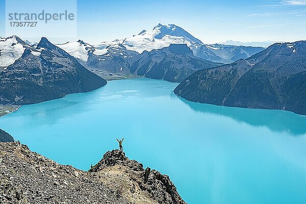 Junger Mann steht auf einem Felsen  streckt die Arme in die Luft  Blick auf Berge und Gletscher mit türkisblauem See Garibaldi Lake  Gipfel Panorama Ridge  Guard Mountain und Deception Peak  Garibaldi Provincial Park  British Columbia  Kanada  Nordamerika