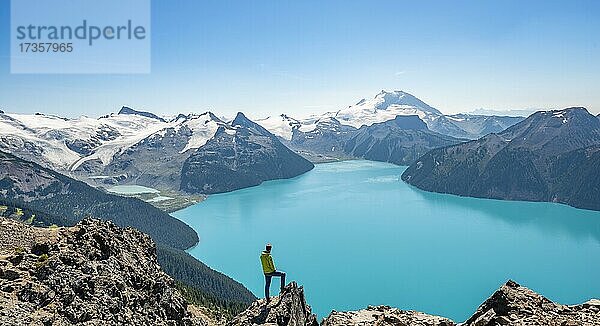 Junger Mann steht auf einem Felsen  blickt in die Ferne  Blick auf Berge und Gletscher mit türkisblauem See Garibaldi Lake  Gipfel Panorama Ridge  Guard Mountain und Deception Peak  Garibaldi Provincial Park  British Columbia  Kanada  Nordamerika