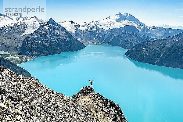 Junger Mann steht auf einem Felsen  streckt die Arme in die Luft  Blick auf Berge und Gletscher mit türkisblauem See Garibaldi Lake  Gipfel Panorama Ridge  Guard Mountain und Deception Peak  Garibaldi Provincial Park  British Columbia  Kanada  Nordamerika