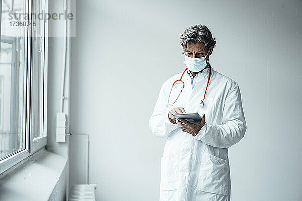 Älterer männlicher Arzt bei der Verwendung eines digitalen Tablets im Krankenhaus während COVID-19