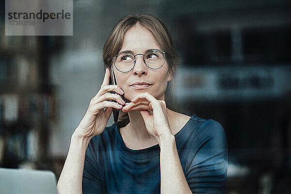 Weibliche Freiberuflerin mit Hand am Kinn  die in einem Café mit ihrem Mobiltelefon spricht