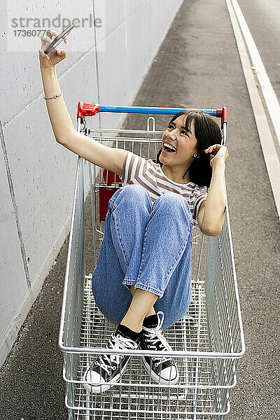 Fröhliche Frau  die ein Selfie mit ihrem Smartphone im Einkaufswagen macht