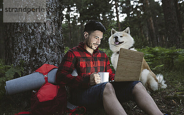 Männlicher Wanderer arbeitet am Laptop  während er bei Sonnenuntergang im Wald neben einem Hund sitzt