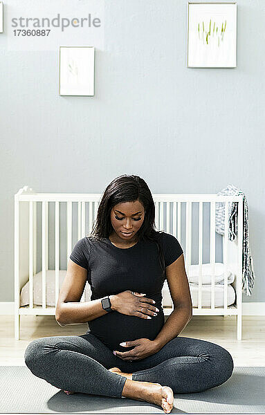 Schwangere Frau  die ihren Bauch berührt  während sie im Schneidersitz auf einer Übungsmatte im Schlafzimmer sitzt