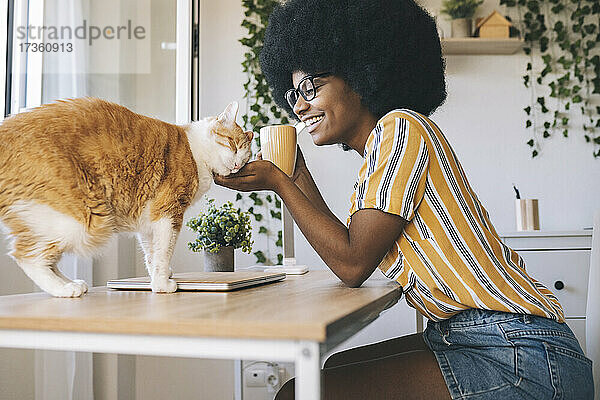 Glückliche Frau mit Kaffeetasse schaut auf Katze auf dem Schreibtisch