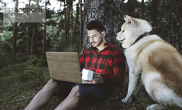 Männlicher Wanderer mit Kaffeetasse und Laptop  während er neben einem Hund im Wald sitzt