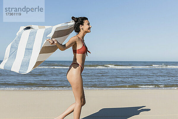 Junge Frau winkt mit dem Handtuch  während sie im Urlaub am Strand läuft