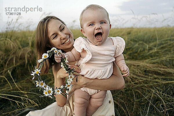 Lächelnde Mutter  die ein fröhliches kleines Mädchen mit einem Blumen-Diadem im Feld hält