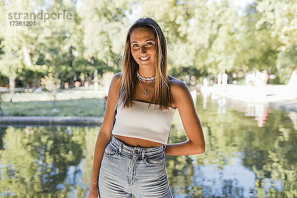 Lächelnde schöne blonde Frau steht vor einem Teich in einem öffentlichen Park