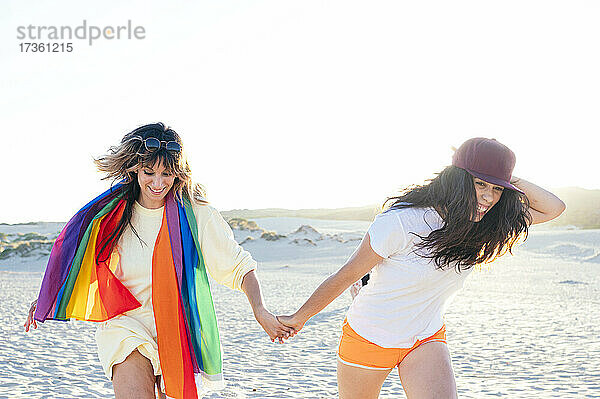 Glückliche Frau mit Regenbogenflagge hält die Hand ihrer Freundin beim Laufen am Strand