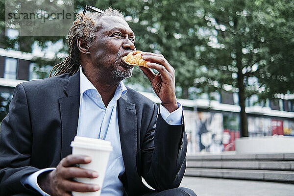Geschäftsmann mit Einwegbecher isst Croissant